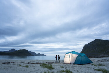 Norway, Lapland, Two people standing by tent on a beach at fjord - KKAF02069