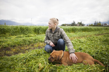 Teenager-Mädchen, das auf einem frisch gemähten Feld kauert und einen Hund streichelt, Chilliwack, British Columbia, Kanada - AURF07316