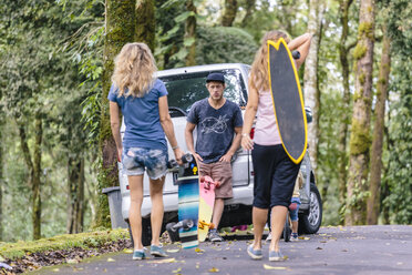 Zwei Frauen mit Skateboards gehen auf einen wartenden männlichen Freund zu, der sich an ein geparktes Auto lehnt, Bali, Indonesien - AURF07421
