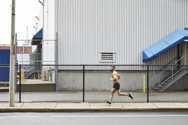 Mann ohne Hemd joggt auf dem Bürgersteig entlang des Zauns eines Industriegebäudes, Boston, Massachusetts, USA - AURF07435