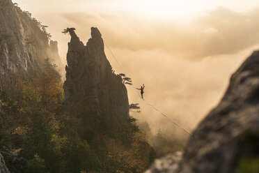 Silhouette eines Mannes, der auf einem zwischen zwei Felsen hängenden Hochseil balanciert, Niederösterreich, Österreich - AURF07500