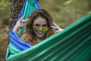 Woman in sunglasses looking playfully at camera while lying in hammock in forest - AURF07595