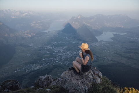 Schweiz, Grosse Mythen, junge Frau beim Wandern auf einem Felsen sitzend bei Sonnenaufgang aus einem Becher trinkend, lizenzfreies Stockfoto