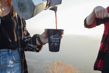 Nahaufnahme einer Frau auf einer Wanderung bei Sonnenaufgang, die einem Freund Kaffee in eine Tasse gießt - LHPF00069