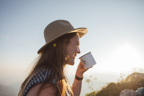 Glückliche junge Frau auf einem Wanderausflug bei Sonnenaufgang mit einer Tasse in der Hand - LHPF00073