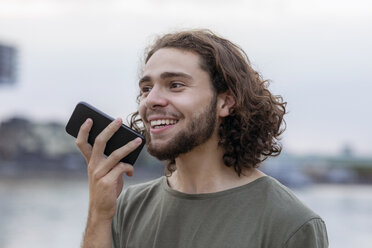 Porträt eines glücklichen jungen Mannes, der sein Smartphone im Freien benutzt - FMKF05253