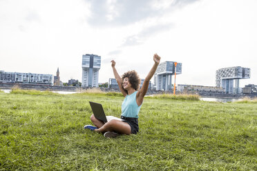 Germany, Cologne, cheering woman sitting on a meadow using laptop - FMKF05254