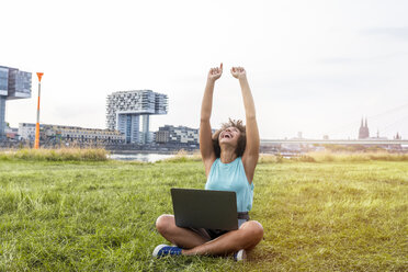 Germany, Cologne, cheering woman sitting on a meadow using laptop - FMKF05255