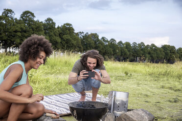 Smiling young man with girlfriend taking a picture of barbecue grill in the nature - FMKF05264