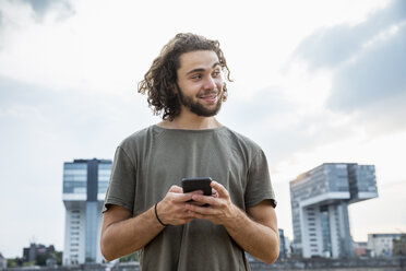 Deutschland, Köln, lächelnder junger Mann hält Handy und schaut zur Seite - FMKF05273