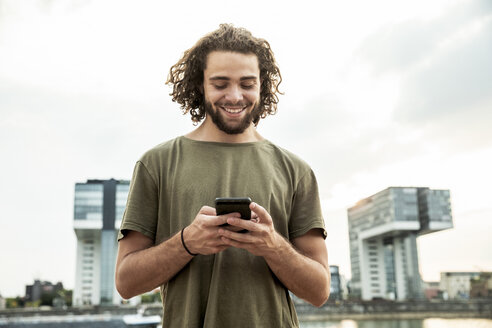 Deutschland, Köln, lächelnder junger Mann schaut auf sein Handy - FMKF05274