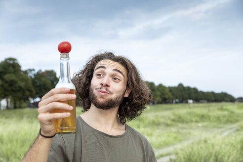 Porträt eines lächelnden jungen Mannes im Freien, der eine Tomate auf einer Bierflasche balanciert - FMKF05280