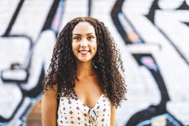 Portrait of smiling young woman with long curly hair at graffiti wall - WPEF00784