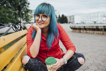 Porträt einer lächelnden jungen Frau mit gefärbten blauen Haaren, die auf einer Bank mit einem Getränk sitzt - VPIF00830