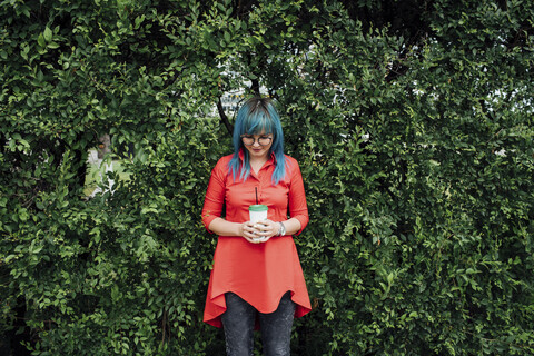 Junge Frau mit gefärbten blauen Haaren steht vor einer Hecke mit Getränk, lizenzfreies Stockfoto