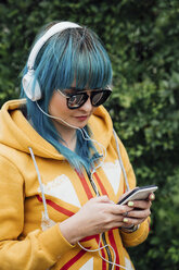 Porträt einer jungen Frau mit gefärbten blauen Haaren, die mit Kopfhörern Musik hört und auf ihr Smartphone schaut - VPIF00840