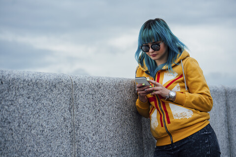 Porträt einer jungen Frau mit gefärbten blauen Haaren, die an der Wand lehnt und auf ihr Handy schaut, lizenzfreies Stockfoto