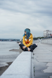 Junge Frau mit gefärbten blauen Haaren sitzt auf einer Mauer und schaut in die Ferne - VPIF00854