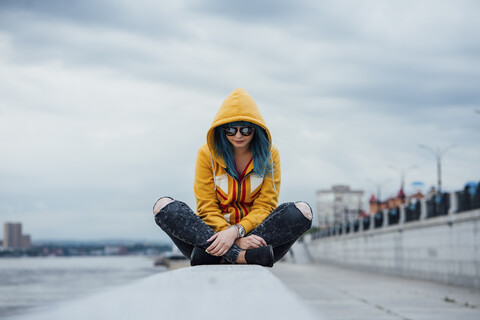 Junge Frau mit gefärbten blauen Haaren sitzt auf einer Mauer und trägt eine modische Kapuzenjacke, lizenzfreies Stockfoto