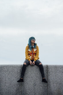 Junge Frau mit gefärbten blauen Haaren sitzt auf einer Mauer und schaut in die Ferne - VPIF00861