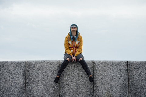 Junge Frau mit gefärbten blauen Haaren, die auf einer Mauer sitzt und nach oben schaut, lizenzfreies Stockfoto