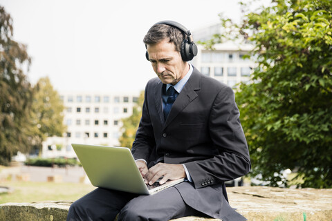 Geschäftsmann im Stadtpark, der Kopfhörer trägt und einen Laptop benutzt, lizenzfreies Stockfoto