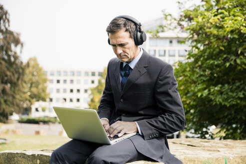 Geschäftsmann im Stadtpark, der Kopfhörer trägt und einen Laptop benutzt - MOEF01379
