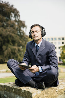 Geschäftsmann im Stadtpark mit Kopfhörern und Tablet in der Hand - MOEF01380