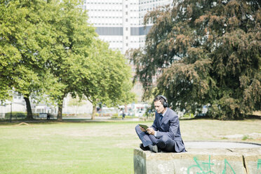 Geschäftsmann im Stadtpark, der Kopfhörer trägt und ein Tablet benutzt - MOEF01399