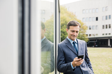 Smiling businessman using cell phone leaning against glass front - MOEF01410