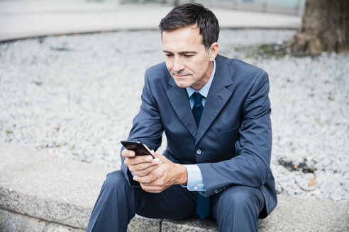 Geschäftsmann sitzt und benutzt ein Mobiltelefon in der Stadt - MOEF01421