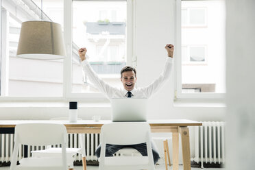 Laughing businessman with laptop on table cheering in office - MOEF01448