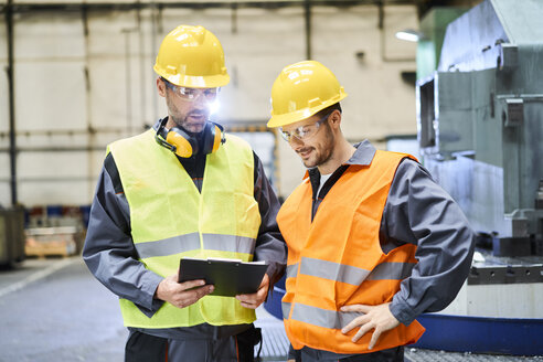 Zwei Männer in Arbeitsschutzkleidung halten ein Klemmbrett und unterhalten sich in einer Fabrik - BSZF00589