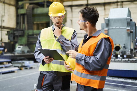 Zwei Männer in Arbeitsschutzkleidung halten ein Klemmbrett und unterhalten sich in einer Fabrik, lizenzfreies Stockfoto