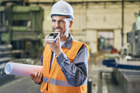 Lächelnder Mann, der Blaupausen hält und ein Handy in einer Fabrik benutzt - BSZF00607