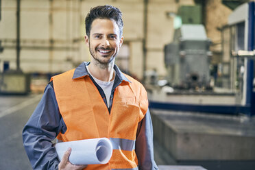 Porträt eines lächelnden Mannes mit Blaupausen in einer Fabrik - BSZF00609