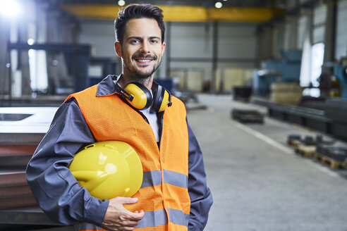 Porträt eines lächelnden Mannes in Arbeitsschutzkleidung in einer Fabrik - BSZF00618
