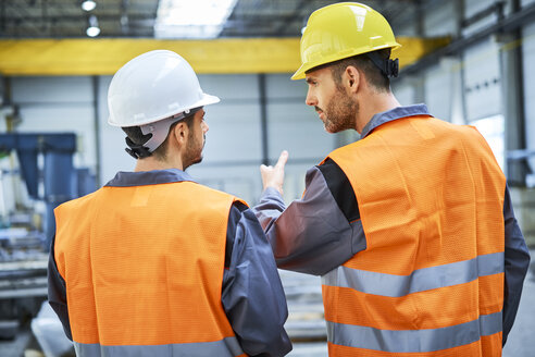 Rückansicht von zwei Männern in Arbeitsschutzkleidung, die sich in einer Fabrik unterhalten - BSZF00628
