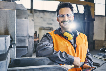 Porträt eines lächelnden Mannes in Arbeitsschutzkleidung mit einem Tablet in einer Fabrik - BSZF00638