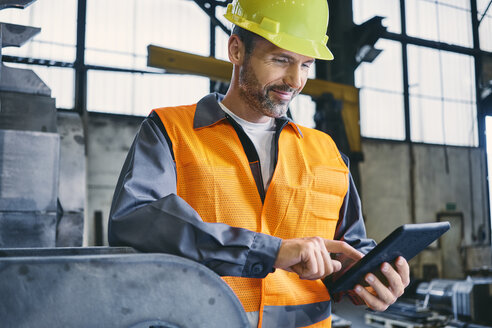 Lächelnder Mann in Arbeitsschutzkleidung bei der Arbeit mit einem Tablet in einer Fabrik - BSZF00642