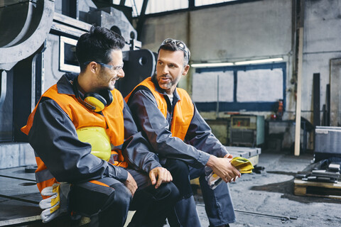 Zwei Männer in Arbeitsschutzkleidung unterhalten sich während einer Pause in einer Fabrik, lizenzfreies Stockfoto