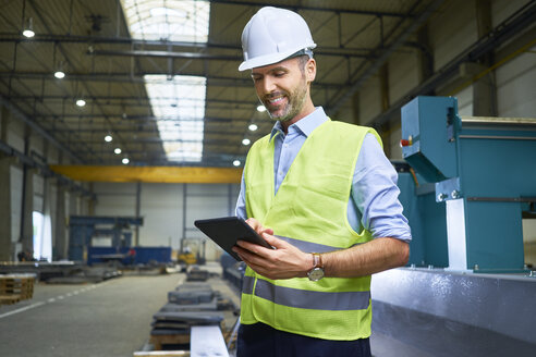 Lächelnder Mann mit Hemd und Sicherheitsweste, der in einer Fabrik ein Tablet benutzt - BSZF00658