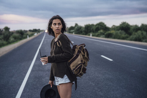 Porträt einer jungen trampenden Frau mit Rucksack und Getränk auf der Fahrbahn stehend - VPIF00903