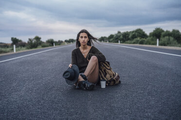 Portrait of hitchhiking young woman with backpack and beverage sitting on lane - VPIF00907