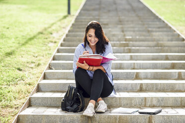 Lächelnder Student, der auf einer Treppe im Freien sitzt und Notizen in ein Notizbuch schreibt - JSMF00450