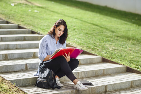 Studentin sitzt auf einer Treppe im Freien und macht sich Notizen in einem Notizbuch - JSMF00451
