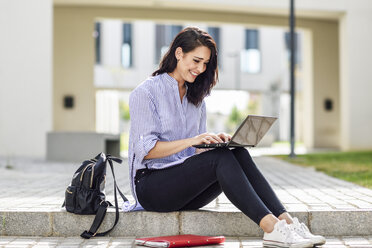 Lächelnder Student, der auf einer Treppe im Freien sitzt und am Laptop arbeitet - JSMF00453