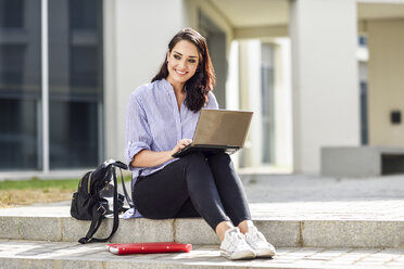 Porträt eines lächelnden Studenten, der auf einer Treppe im Freien sitzt und an einem Laptop arbeitet - JSMF00455