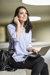 Porträt einer lächelnden Studentin mit Laptop, die Kopfhörer aufsetzt - JSMF00460
