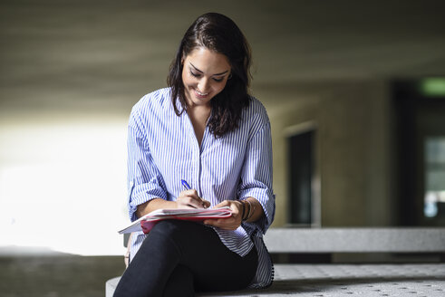 Lächelnde Studentin sitzt auf einer Bank im Freien und macht sich Notizen in einem Notizbuch - JSMF00461